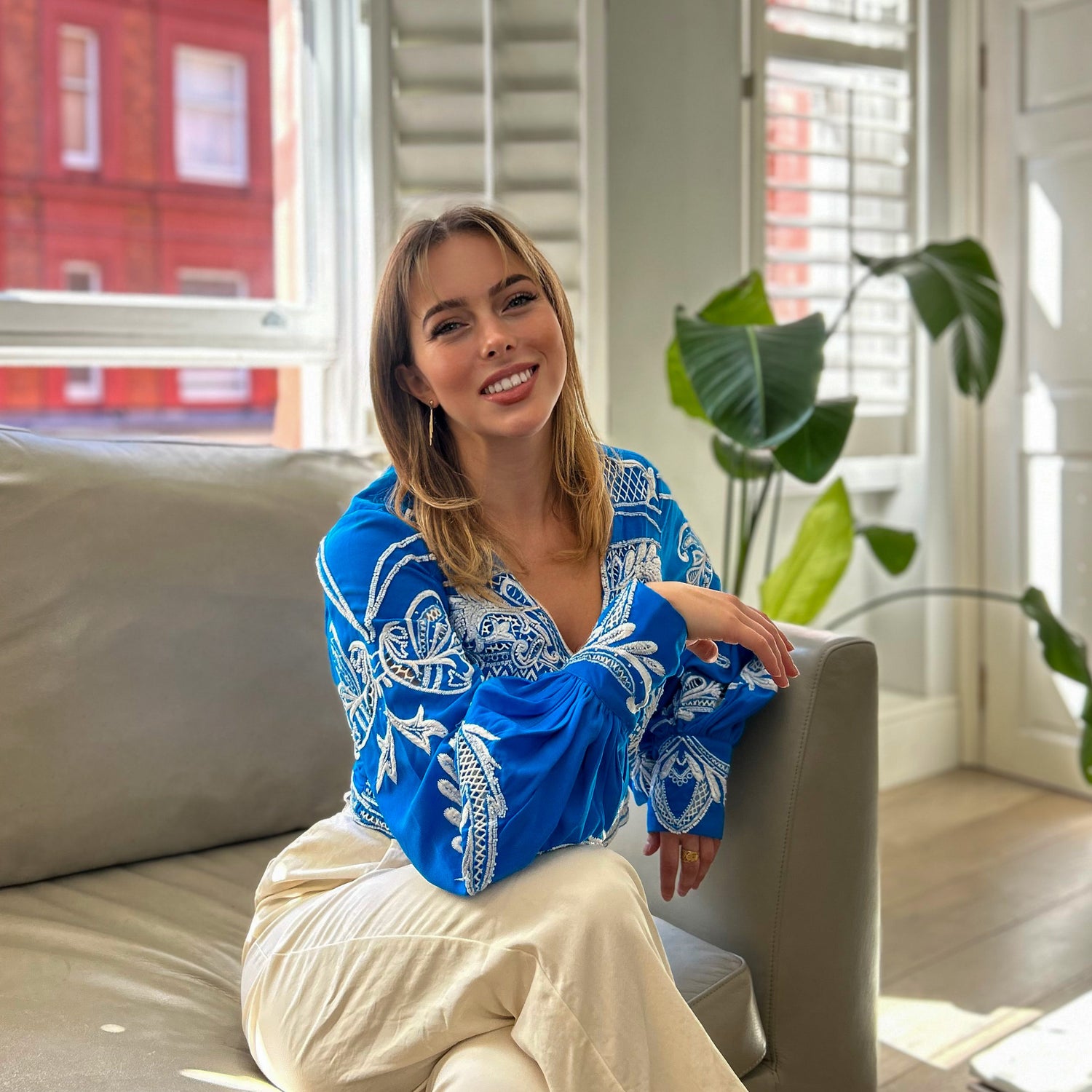 Woman in a blue embroidered top sitting on a couch in a bright room with plants.