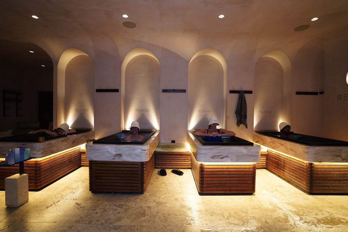 A spa relaxation room with four marble and wood plunge pools, where people are reclining under warm ambient lighting in an arched stone interior.