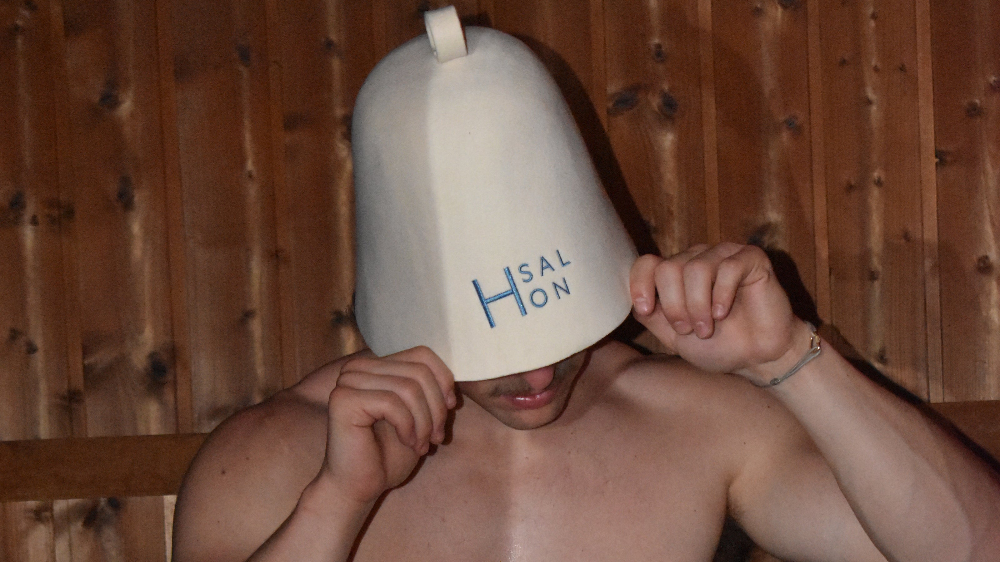 A person in a sauna wearing a white ceramic Hålön pot holder with blue lettering, set against a wooden wall.