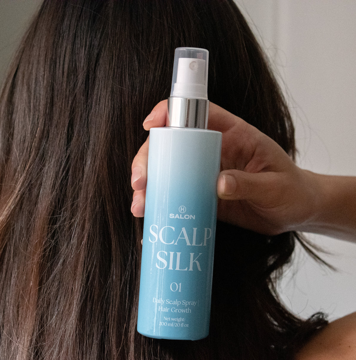 Person holding a bottle of  H Salon Scalp Silk spray with long hair in the background